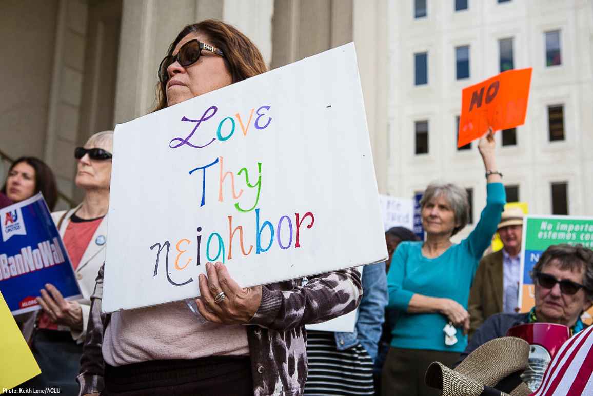 A Latina woman holding a sign that says "Love Thy Neighbor"