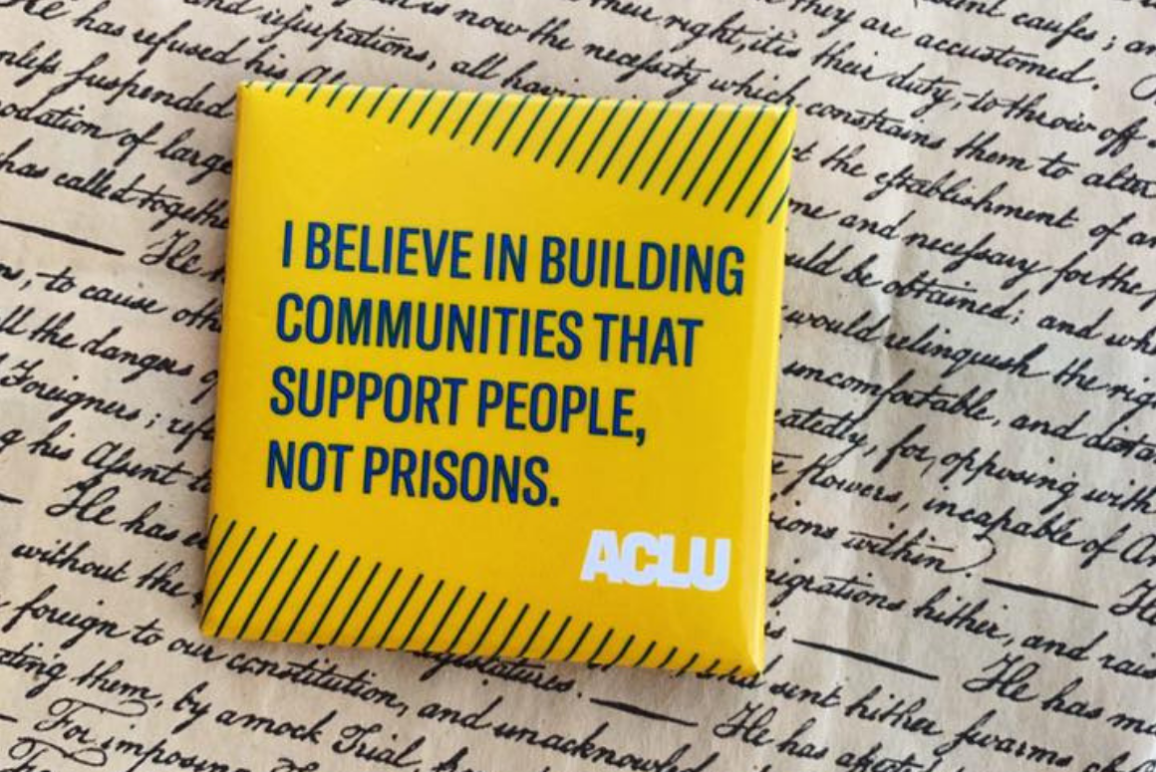 a square yellow button with green text that says "I believe in building communities that support people, not prisons" against a background of cursive writing on parchment paper.