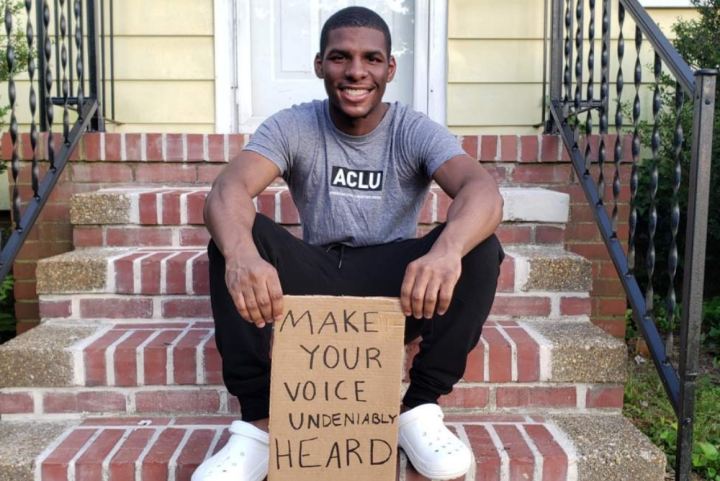 a photo of our intern Alton Coston sitting on the steps in front of his house, holding a cardboard sign that says "make your voice undeniably heard"