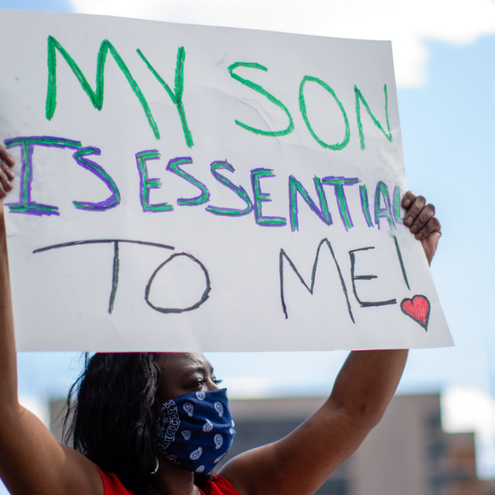 a black woman wearing mask and holding a sign that says "my son is essential to me"