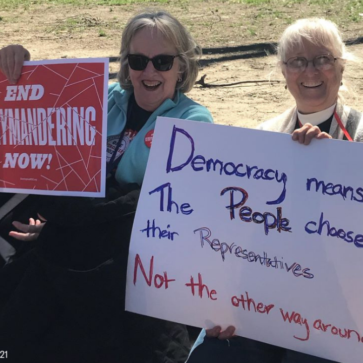 two protesters holding signs that say "End gerrymandering now" and "Democracy means the people choose their reps, not the other way around"